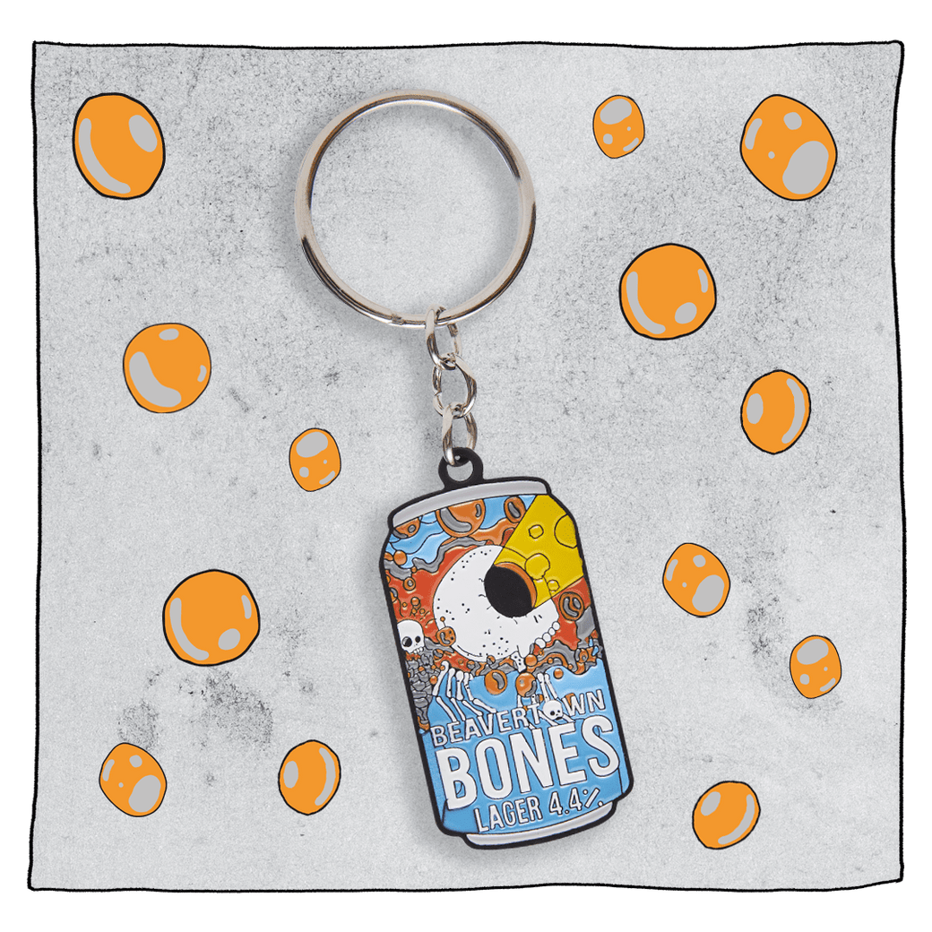 Beavertown Brewery Bones Keyring. Keyring is small flat replica of the Bones blue and orange can with white skull with skeleton hands. Skull only has one large eye socket which is exerting a yellow lazer beam. Grey background with orange spots around keyring.