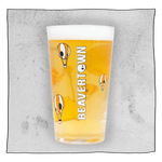 Neck Oil pint glass filled with beer in front of a light grey background. Glass has many orange and white skull hot air balloons and white Beavertown Logo.