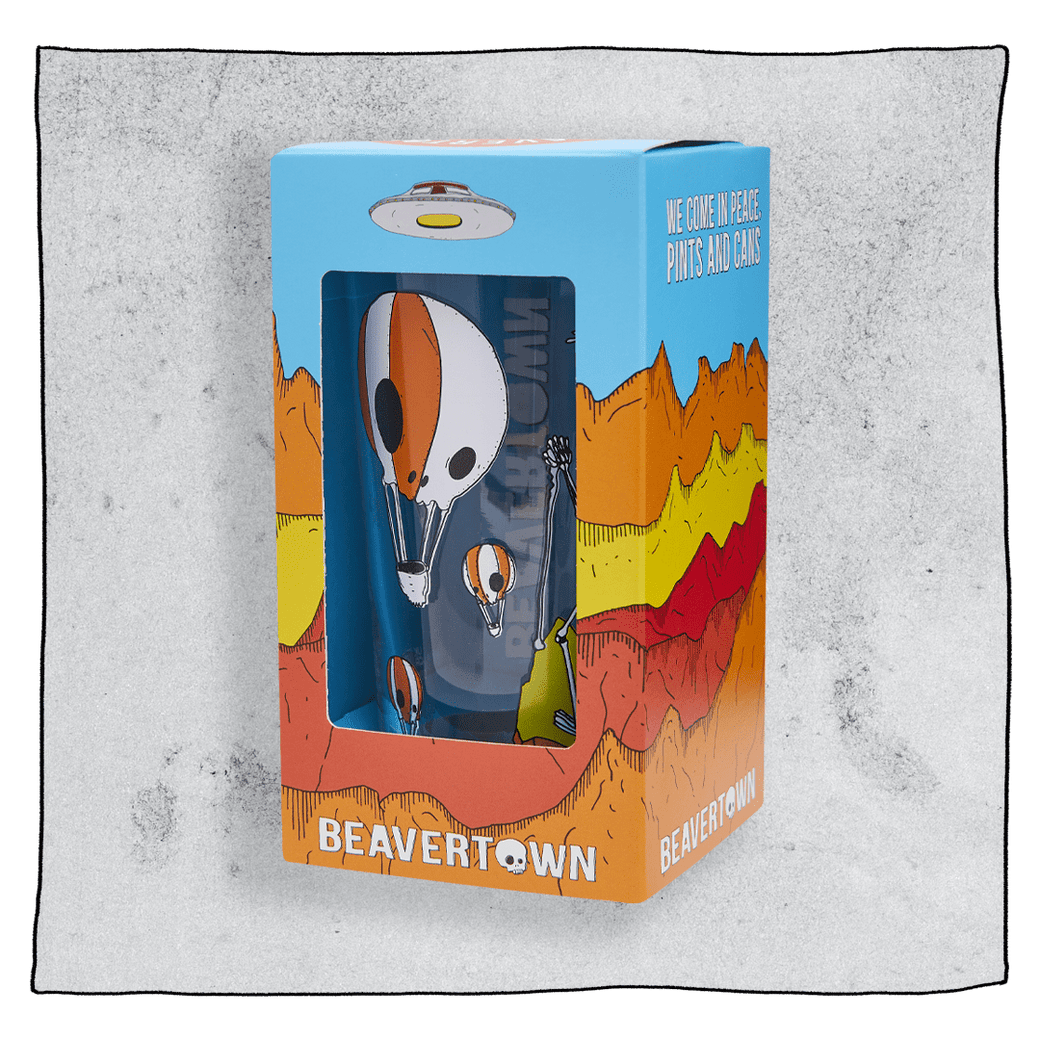 Beavertown Neck Oil pint glass inside an orange and blue box in front of a grey background. Box has artwork depicting a white UFO in an orange, yellow and red canyon with a blue sky. Neck Oil pint glass is visible in middle of box and is clear glass with orange and white skull hot air balloons. Grey background.