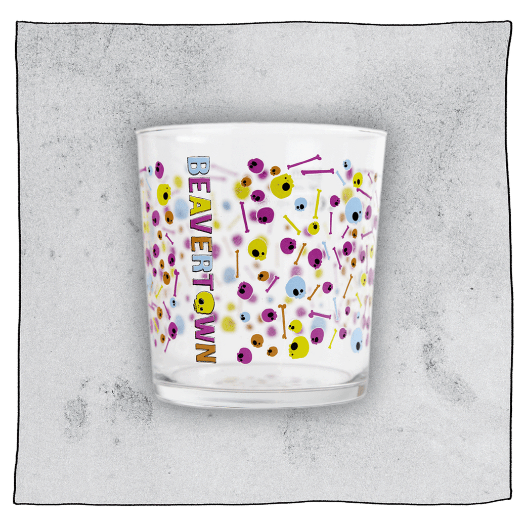 Empty Psychedelic tumbler glass in front of a grey background. Glass is clear with colourful skulls and bones scattered all around the glass.