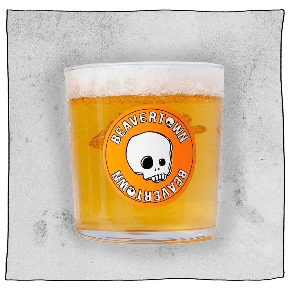 Beavertown Half pint glass filled with beer in front of a grey background. Beer glass is clear glass but contains an orange Beavertown logo with a white skull.