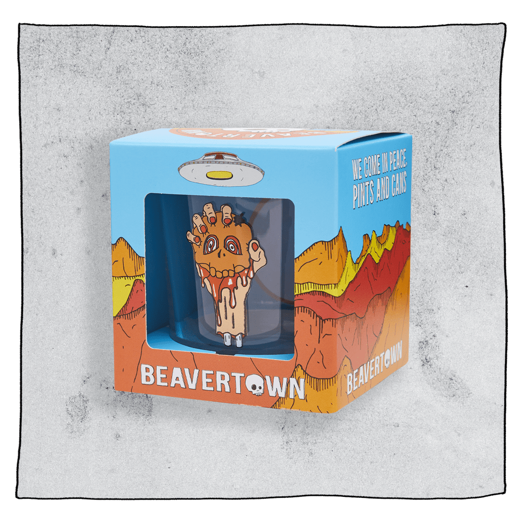 Beavertown Blood E'll Half pint glass inside an orange and blue cubed box in front of a grey background. Box has artwork depicting a white UFO in an orange, yellow and red canyon with a blue sky. Blood E'll half pint glass is visible in middle of box and is clear glass but contains an image of a bloody severed arm with orange fingernails clenching a small orange skull.