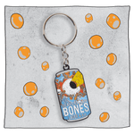 Beavertown Brewery Bones Keyring. Keyring is small flat replica of the Bones blue and orange can with white skull with skeleton hands. Skull only has one large eye socket which is exerting a yellow lazer beam. Grey background with orange spots around keyring.