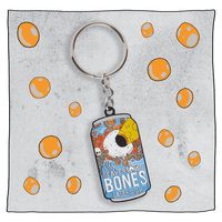 Beavertown Brewery Bones Keyring. Keyring is small flat replica of the Bones blue and orange can with white skull with skeleton hands. Skull only has one large eye socket which is exerting a yellow lazer beam. Grey background with orange spots around keyring.