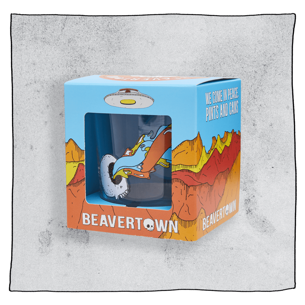 Beavertown Bones tumbler glass inside an orange and blue cubed box in front of a grey background. Box has artwork depicting a white UFO in an orange, yellow and red canyon with a blue sky. Bones tumbler glass is visible in middle of box and is clear glass with logo of white skull with one eye socket shooting yellow beam amongst orange and blue streaks. Grey background.