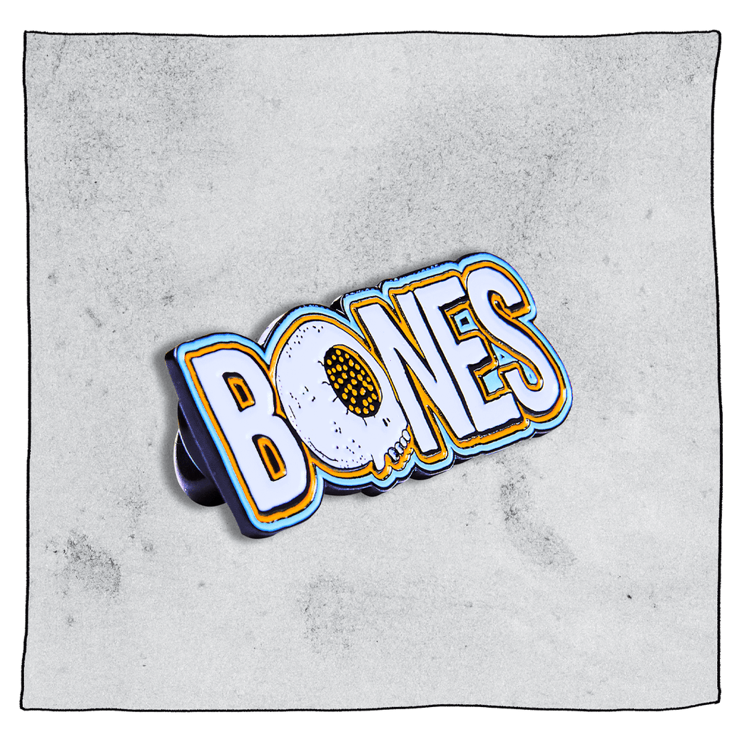 Bones Pin Badge. Left side angled view. White bones badge with orange and blue outline of text and white skull with orange eye instead the letter 'O'. Grey background.