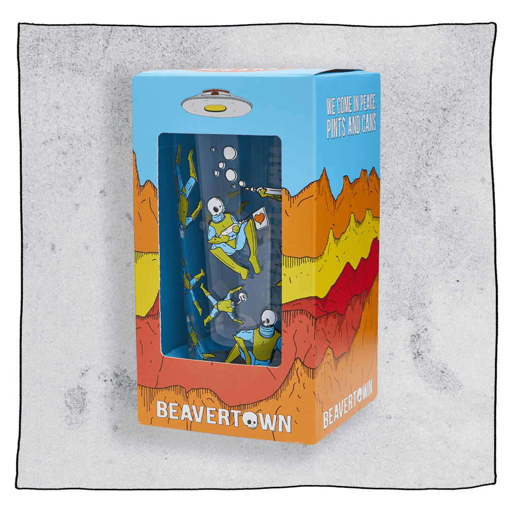 Beavertown Gamma Ray pint glass inside an orange and blue box in front of a grey background. Box has artwork depicting a white UFO in an orange, yellow and red canyon with a blue sky. Gamma Ray pint glass is visible in middle of box and is clear glass with blue and yellow gamma ray men around the glass. Grey background.