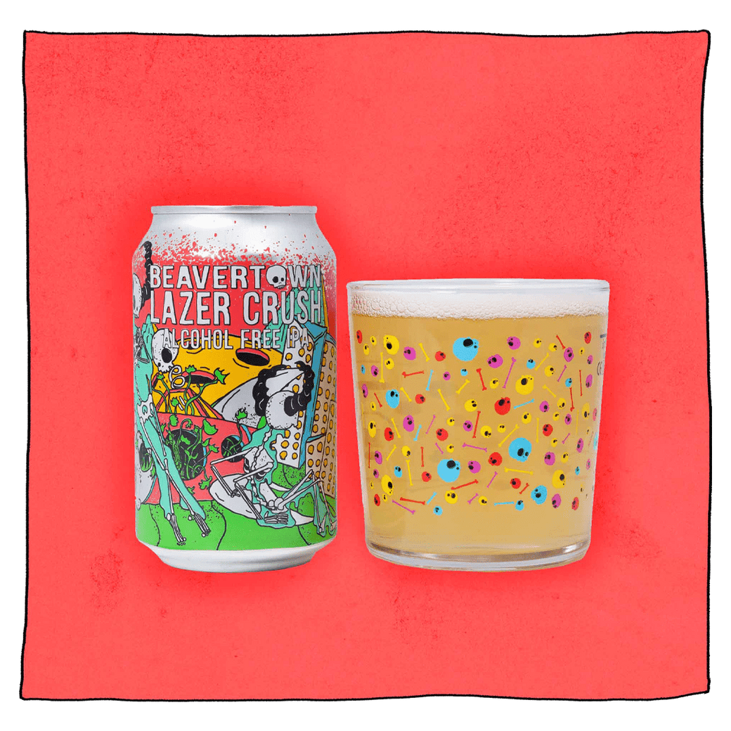 Beavertown Brewery Lazer Crush - Alcohol Free IPA and Beavertown Psychedelic Skull Tumbler glass filled with beer in front of a pink background. Beer glass is clear glass that has red, blue, pink and yellow skulls and bones speckled all over.