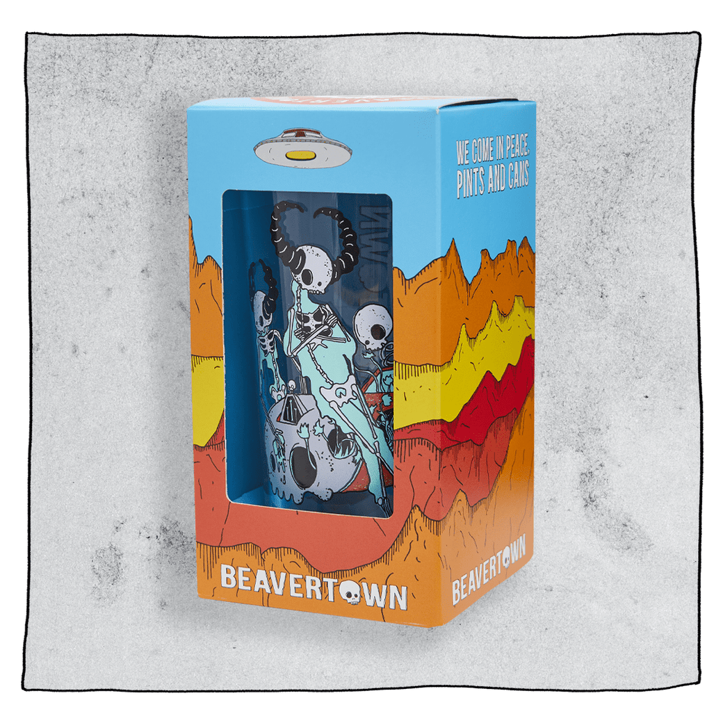 Beavertown Lazer Crush pint glass inside an orange and blue box in front of a grey background. Box has artwork depicting a white UFO in an orange, yellow and red canyon with a blue sky. Lazer Crush pint glass is visible in middle of box and is clear glass with white skeletons with black horns.