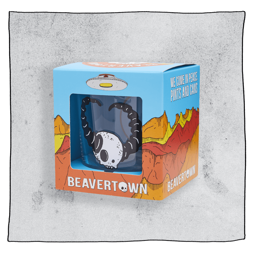 Beavertown Lazer Crush Tumbler glass inside an orange and blue box in front of a grey background. Box has artwork depicting a white UFO in an orange, yellow and red canyon with a blue sky. Lazer Crush Tumbler glass is visible in middle of box and is clear glass with white skull with black horns.