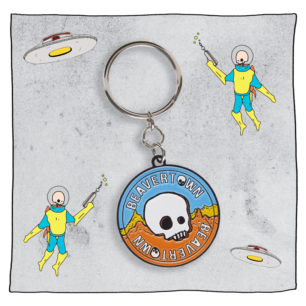 Beavertown Brewery circle metal keyring of the Beavertown logo - Beavertown written in white twice around the edge of the circle with a skull in the middle in front of a blue sky and yellow and orange mountains.