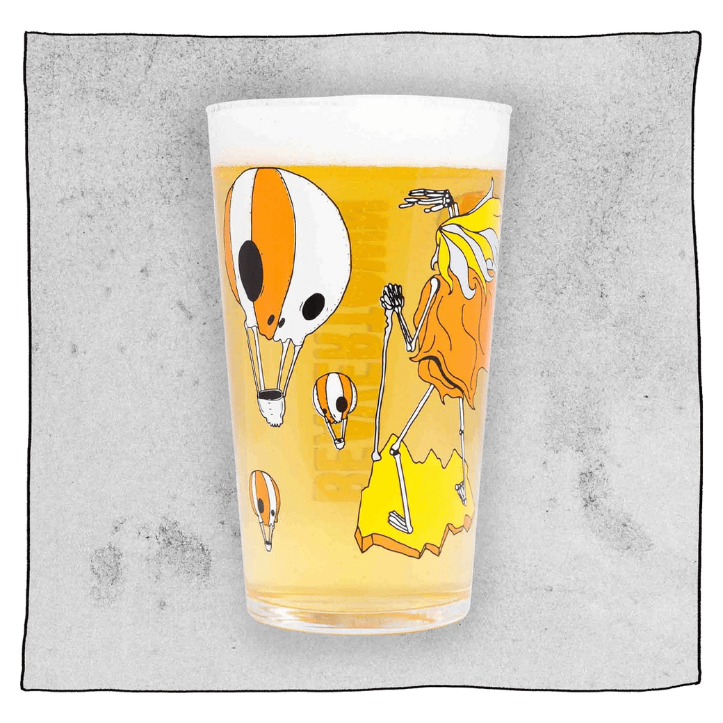Neck Oil pint glass filled with beer in front of a light grey background. Glass has many orange and white skull hot air balloons.