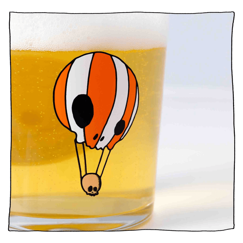 Zoomed in shot of Neck Oil tumbler glass filled with beer in front of a light grey background. Glass is clear with an orange and white skull hot air balloon.
