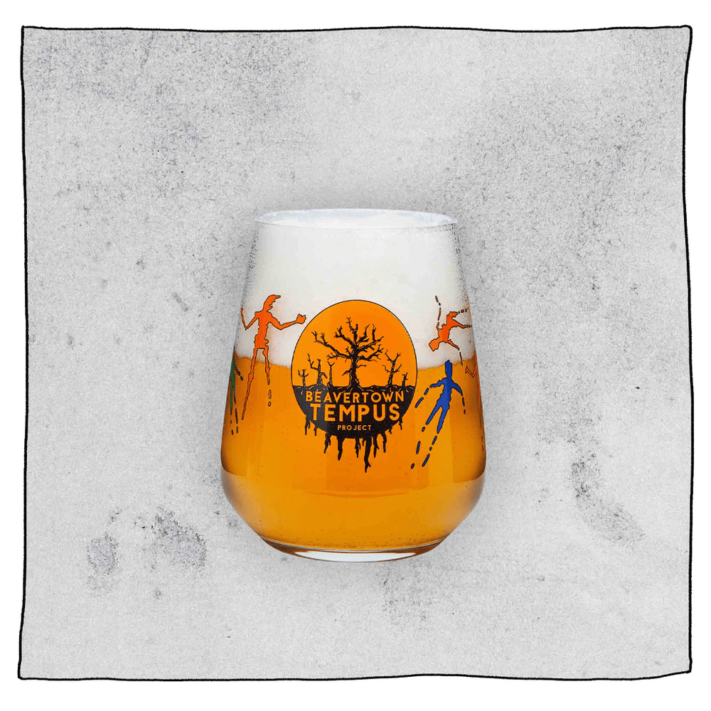 Tempus Tumbler glass filled with beer in front a grey background. Glass has orange and black  dead tree Tempus logo.